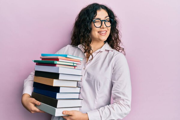 Young brunette woman with curly hair holding a pile of books winking looking at the camera with sexy expression, cheerful and happy face. 