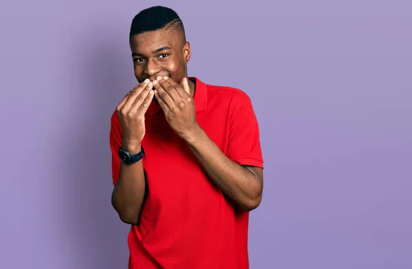 Young african american man wearing casual red t shirt laughing and embarrassed giggle covering mouth with hands, gossip and scandal concept