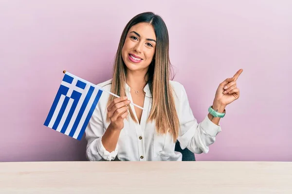 Jovem Hispânica Segurando Bandeira Grega Sentada Mesa Sorrindo Feliz Apontando — Fotografia de Stock