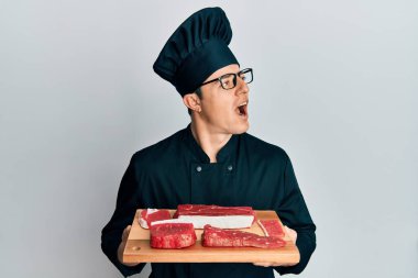 Yakışıklı genç adam, şef üniforması giyiyor, elinde çiğ etle kızgın ve kızgın çığlıklar atıyor, öfkeyle bağırıyor. öfke ve agresif konsept. 