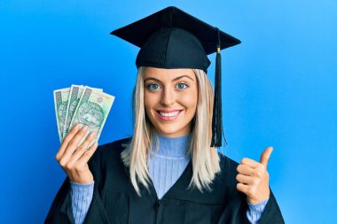 Mezuniyet şapkası ve tören cübbesi takan güzel sarışın kadın elinde cila zloti, gülümseyen mutlu ve olumlu bir şekilde başparmağıyla mükemmel ve onay işareti yapıyor. 