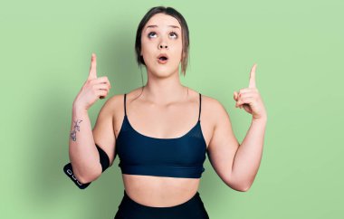 Spor kıyafetleri giyen genç İspanyol kadın şaşırmış ve şaşırmış bir şekilde yukarı bakıyor ve parmaklarıyla işaret ediyor ve kolunu kaldırıyor.. 