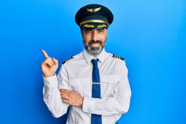 Orta yaşlı, sakallı, gri saçlı, yüzünde kocaman bir gülümseme olan uçak pilotu üniforması giyen, kameraya el ve parmağıyla bakan bir adam.. 