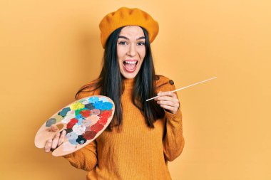 Elinde boya fırçası ve palet tutan genç esmer kadın çılgın kutlama beresi takıyor ve başarı için hayrete düşüyor açık gözlerle heyecanla haykırıyor. 