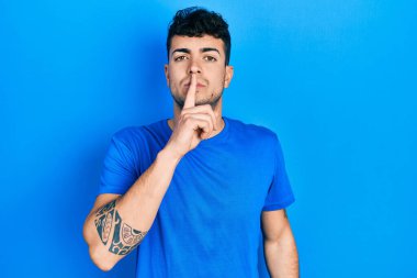 Latin asıllı genç bir adam günlük mavi tişört giyiyor ve dudakları parmaklarıyla sessiz olmak istiyor. sessizlik ve gizli konsept. 