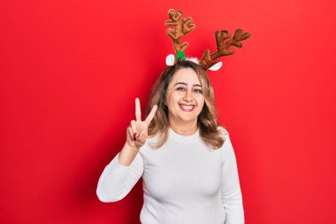 Orta yaşlı beyaz kadın şirin Noel geyiği boynuzları takıyor. İki numarayı gösterip işaret ederken kendine güvenen ve mutlu bir şekilde gülümsüyor.. 