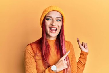 Genç beyaz kadın yün şapka takıyor gülümsüyor ve kameraya bakıyor. İki eli ve parmağı yana bakıyor.. 