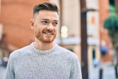 Genç beyaz adam gülümseyerek şehirde mutlu mesut duruyor..