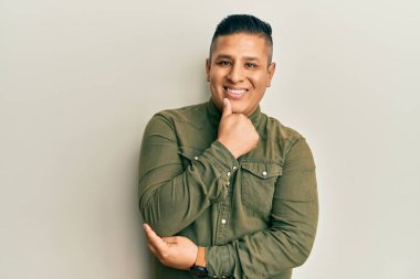 Gündelik kıyafetler giyen genç latin adam kameraya bakıyor kollarını kavuşturup gülümsüyor ve çenesini kaldırıyor. Olumlu düşünmek. 