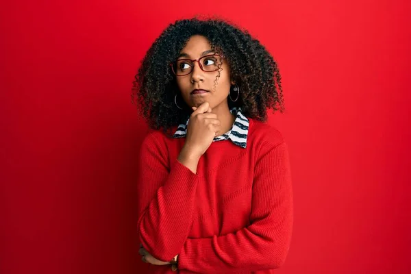 Beautiful african american woman with afro hair wearing sweater and glasses with hand on chin thinking about question, pensive expression. smiling and thoughtful face. doubt concept.