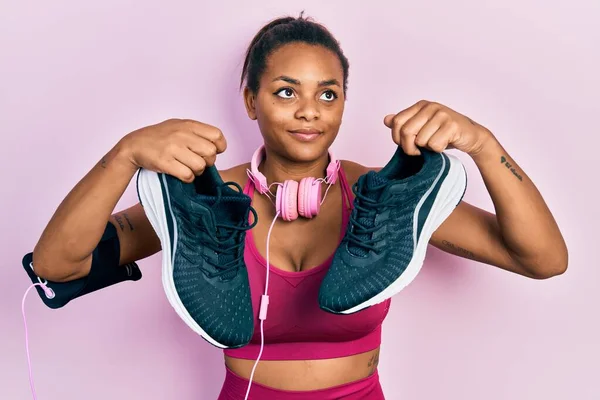 年轻的非洲裔美国女孩穿着运动服 戴着耳机 拿着运动鞋 面带微笑地看着旁边 目不转睛地看着外面想 — 图库照片