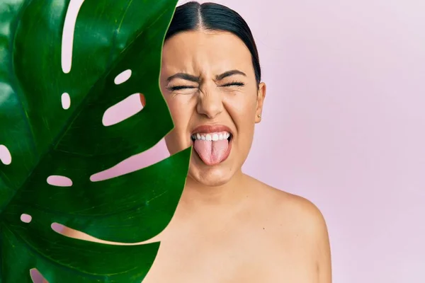 Beautiful brunette woman holding green plant leaf close to beautiful face sticking tongue out happy with funny expression.