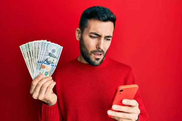 Young Hispanic Man Using Smartphone Holding Usa Dollars Banknotes Clueless — Stock Photo, Image