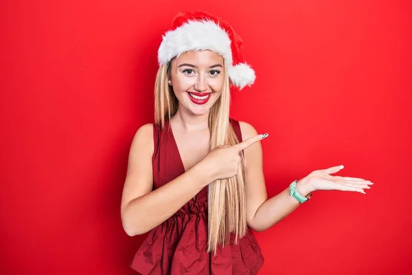 若いですブロンド女の子身に着けていますクリスマスの帽子驚きと笑顔にカメラに提示しながら手とポインティングとともに指 — ストック写真
