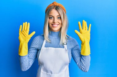 Güzel sarışın kadın daha temiz bir önlük ve eldiven giyer ve parmaklarını on numaraya gösterir ve gösterir. Bu arada kendinden emin ve mutlu gülümser.. 