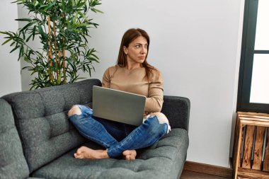 Orta Doğu 'lu üst düzey bir kadın evinde dizüstü bilgisayar kullanıyor. Yan tarafa bakıyor, rahat bir görünüm takınıyor. Kendine güvenen bir gülümsemesi var.. 