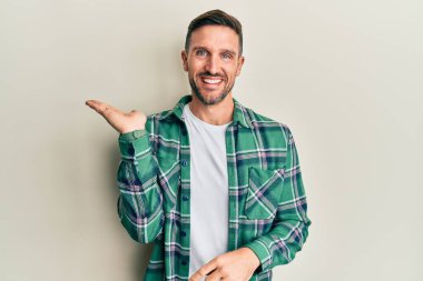 Sakallı, sıradan giyinen, neşeli gülümseyen, eli avucunda kameraya bakan yakışıklı bir adam.. 