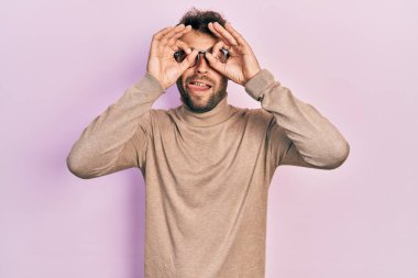 Balıkçı yaka kazak giyen ve gözlük takan yakışıklı bir adam dürbün gibi jest yapıyor. Dürbün dilini çıkarıyor, gözler parmaklarından bakıyor. çılgın ifade. 