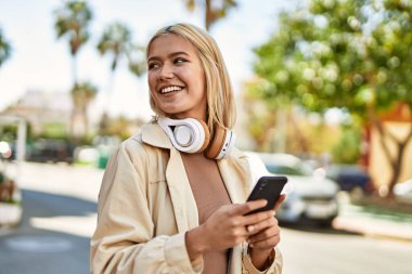 Şehirde akıllı telefon ve kulaklık kullanarak mutlu gülümseyen genç sarışın kız..