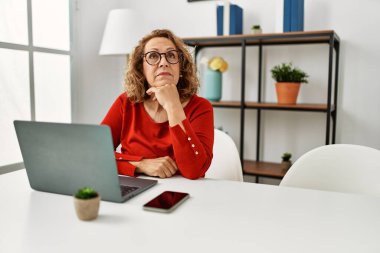 Orta yaşlı, beyaz bir kadın evde dizüstü bilgisayar kullanıyor. Ciddi bir yüz ifadesi var. Elleri çenesinde soru sormayı düşünüyor. 