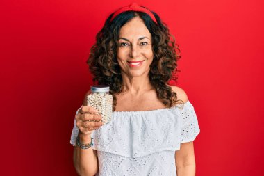 Orta yaşlı İspanyol bir kadın elinde fasulye kavanozu tutarken pozitif ve mutlu görünüyor. Kendine güvenen bir gülümsemeyle dişlerini gösteriyor. 