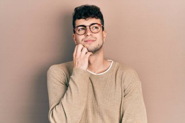 Gündelik kıyafetler giyen ve eli çenesinde gözlüklü soru soran genç İspanyol adam, düşünceli yüz ifadesi. Gülümseyen ve düşünceli bir yüz. şüphe kavramı. 