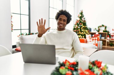Genç Afro-Amerikan adam dizüstü bilgisayar kullanıyor Noel ağacının yanında oturuyor ve beş numarayı işaret ediyor kendinden emin ve mutlu bir şekilde gülümsüyor.. 
