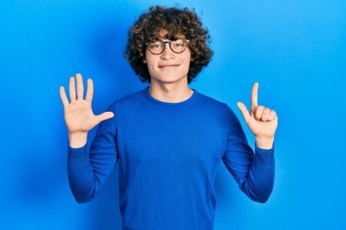 Yakışıklı genç adam günlük kıyafetler giyiyor ve gözlük takıyor. Yedi numaralı parmaklarıyla gösteriyor ve işaret ediyor. Kendinden emin ve mutlu bir şekilde gülümsüyor.. 