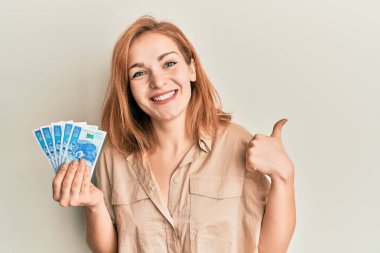 Kafkasyalı genç bir kadın 50 Polonya parası tutuyor mutlu ve olumlu gülümsüyor, başparmağıyla mükemmel ve onay işareti yapıyor. 