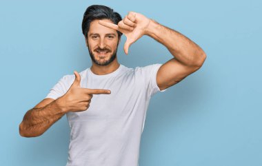 Gündelik beyaz tişört giyen genç İspanyol adam gülümseyerek elleri ve parmakları gülen yüzle çerçeve yapıyor. yaratıcılık ve fotoğrafçılık kavramı. 