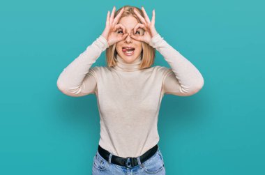 Sıradan kıyafetler giyen genç sarışın bir kadın dürbün gibi jest yapıyor, gözleri parmaklarından bakıyor. çılgın ifade. 