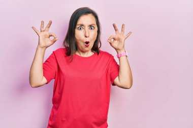 Orta yaşlı İspanyol kadın sıradan kıyafetler giyiyor. Şaşırmış ve şok olmuş görünüyor. Onaylama sembolünü parmaklarıyla yapıyor. çılgın ifade 