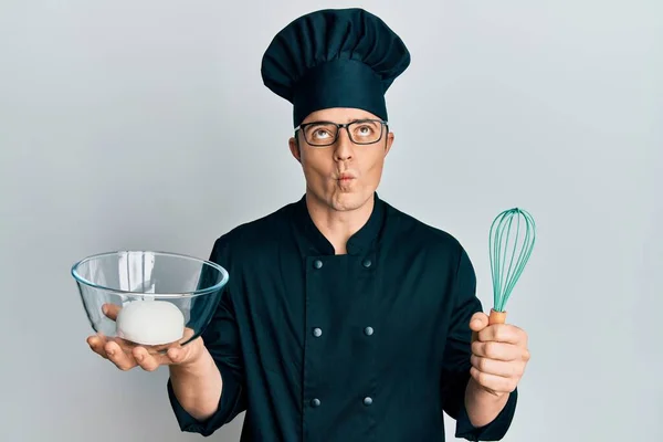 Handsome young man holding bread dough and blender making fish face with mouth and squinting eyes, crazy and comical.