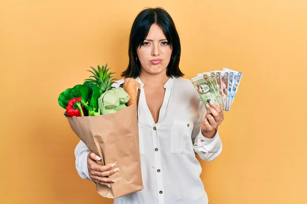 年轻的慌张女人拿着食品杂货袋和秘鲁索尔钞票 怀疑和紧张 皱着眉头因为问题而不高兴 消极的人 — 图库照片