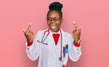 Young african american woman wearing doctor uniform and stethoscope showing middle finger doing fuck you bad expression, provocation and rude attitude. screaming excited 