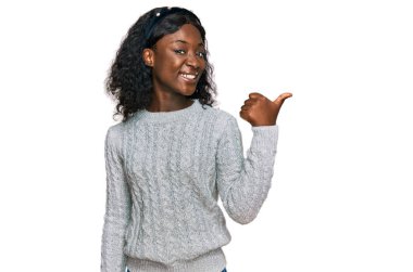 Güzel Afrikalı genç bir kadın, günlük kışlık süveter giyiyor, gülümsüyor, gülümsüyor ve başparmağıyla yan tarafı işaret ediyor.. 