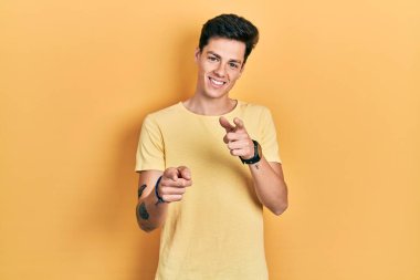 Gündelik sarı tişörtlü genç İspanyol adam mutlu ve komik yüzlü parmaklarını kameraya doğrultuyor. iyi enerji ve titreşimler. 
