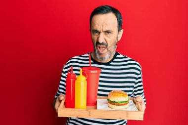 Orta yaşlı İspanyol bir adam lezzetli klasik bir burger ve soda yiyor habersiz ve kafası karışmış bir ifade. şüphe kavramı. 