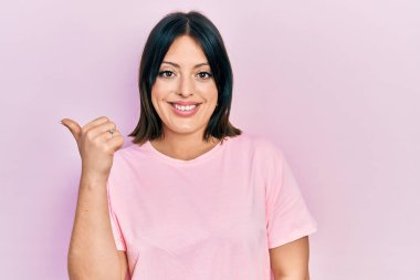 Gündelik pembe tişört giyen, gülümseyen, gülümseyen ve başparmağıyla yan tarafı gösteren genç İspanyol kadın.. 