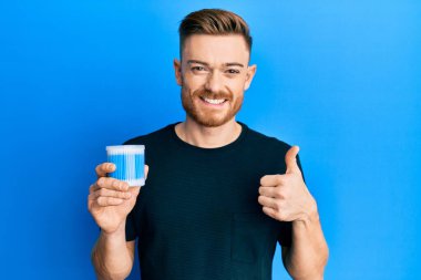 Kulak kiri temizleyici tutan genç kızıl saçlı adam mutlu ve pozitif gülümsüyor, başparmağıyla mükemmel ve onay işareti yapıyor. 