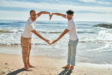 Genç eşcinsel çift gülümseyerek kumsalda kollarıyla kalp sembolü yapıyor..