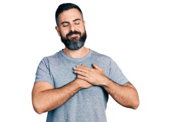 Sakallı, sıradan gri tişörtlü, göğsünde elleriyle gülümseyen, yüzünde minnet ifadesi olan İspanyol bir adam. sağlık kavramı. 