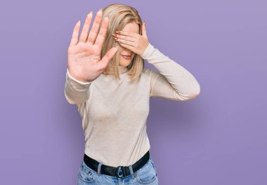 Genç sarışın kadın gündelik kıyafetler giyip gözleri elleriyle kapatıyor ve üzgün ve korku ifadeleriyle jest yapıyor. utangaç ve olumsuz kavram. 