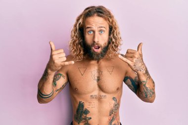 Sakallı, uzun saçlı, gömleksiz, dövmeleri olmayan, Shaka işareti yapan yakışıklı bir adam. Korkmuş ve şaşkınlık içinde, şaşkınlık içinde bir ifade, korku ve heyecan dolu bir yüz.. 
