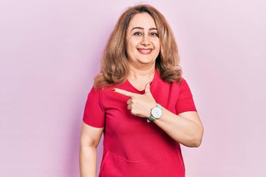 Orta yaşlı, beyaz, günlük kıyafetler giyen, neşeli, yüzünde bir gülümseme olan, mutlu ve doğal bir ifadeyle elini ve parmağını yan tarafına gösteren bir kadın. 