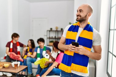 Bir grup İspanyol genç arkadaş futbol maçını izliyor ve destekliyor. Mutlu gülümseyen ve akıllı telefon kullanan bir adam..