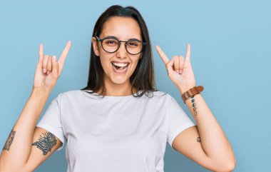 Günlük beyaz tişört giyen genç İspanyol kadın çılgın bir ifadeyle bağırıyor elleri havada rock sembolü yapıyor. Müzik yıldızı. ağır müzik konsepti. 