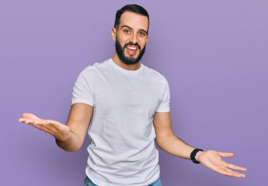 Sakallı genç adam, günlük beyaz tişört giyiyor. Neşeli gülümsüyor, kollarını açıyor. Hoş geldin, olumlu ve kendinden emin selamlar. 