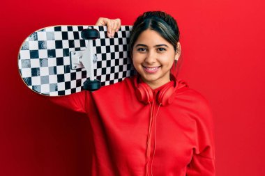 Genç Latin kadın kaykay tutuyor, pozitif ve mutlu görünüyor. Duruyor ve gülümsüyor, kendine güvenen bir gülümsemeyle dişlerini gösteriyor. 