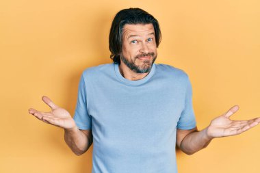 Orta yaşlı, sıradan kıyafetler giyen, elleri ve kolları havada, kafası karışmış bir beyaz adam. şüphe kavramı. 